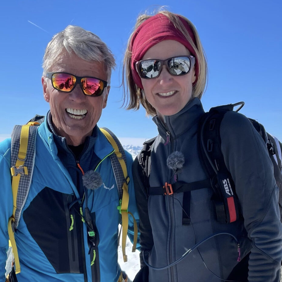 ein sportlicher älterer Herr (Peter Habeler) und eine blonde Frau (Margit Atzler), beide mit Sonnenbrille und Ansteckmikrofonen. Im Hintergrund Berge und ein blauer Himmel.