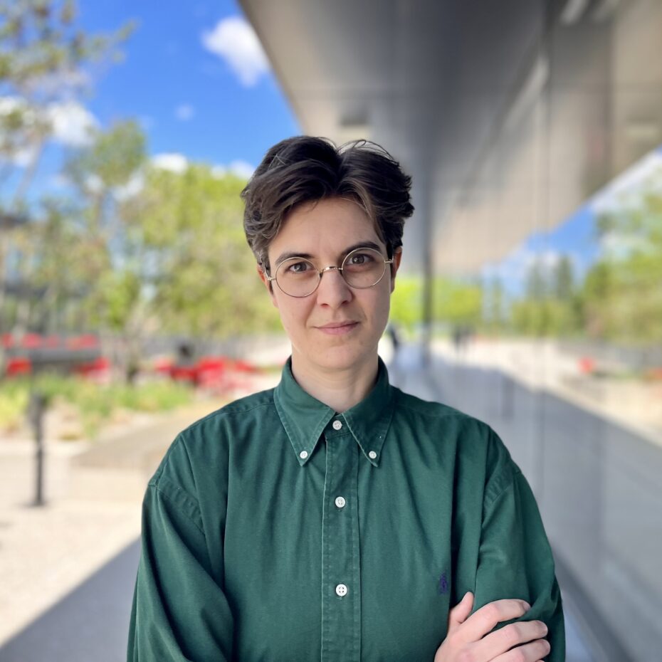 eine Frau mit kurzen Haaren und Brille, in einem grünen Hemd blickt in die Kamera. Im Hintergund spiegeln sich die Bäume entlang einer Glasfront .