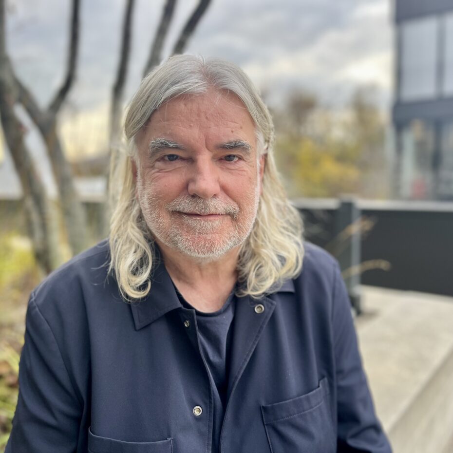 ein Mann mit schulterlangen weißen Haaren und Dreitagesbart blick in die Kamera. Im Hintergrund eine urbane Herbstlandschaft mit kargen Bäumen und modernen Glas- und Betonelementen