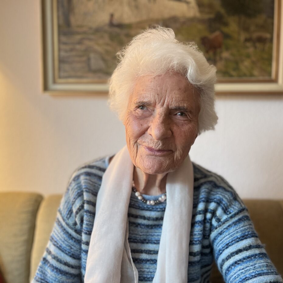 eine weißhaarige Frau mit weißem Schal lächelt in die Kamera. Im Hintergrund ein Bild über dem Sofa, auf dem sie sitzt.