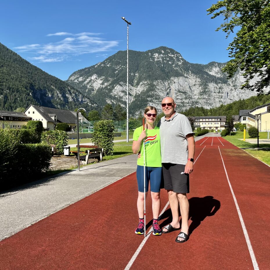 Ein Mann mit Glatze und Sonnenbrille und eine junge Frau mit Brille und Blindenstock stehen auf einer Laufbahn. Im Hintergrund sind weitere Sportanlagen zu erkennen und hohe Berge.