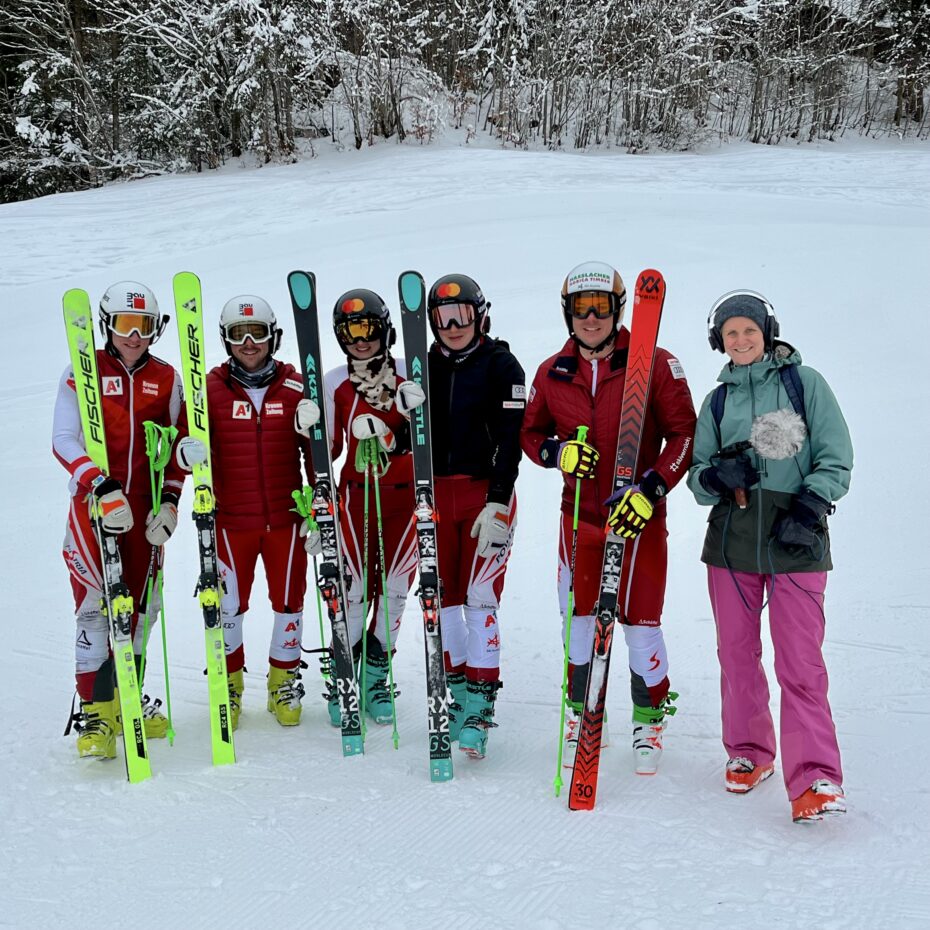 5 Skirennläufer:innen stehen mit ihren Skiern in der Hand und lachen in die Kamera. Daneben eine Frau mit Mikrofon und Kopfhöreren.