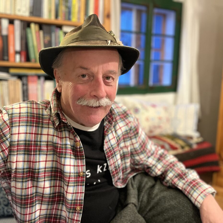 ein Mann mit grauem Schnautzer, kariertem Hemd über einem schwarzen T-Shirt und Filzhut sitzt in der Stube vor einem Bücherregal und Fenster. Er lacht verschmitzt in die Kamera.