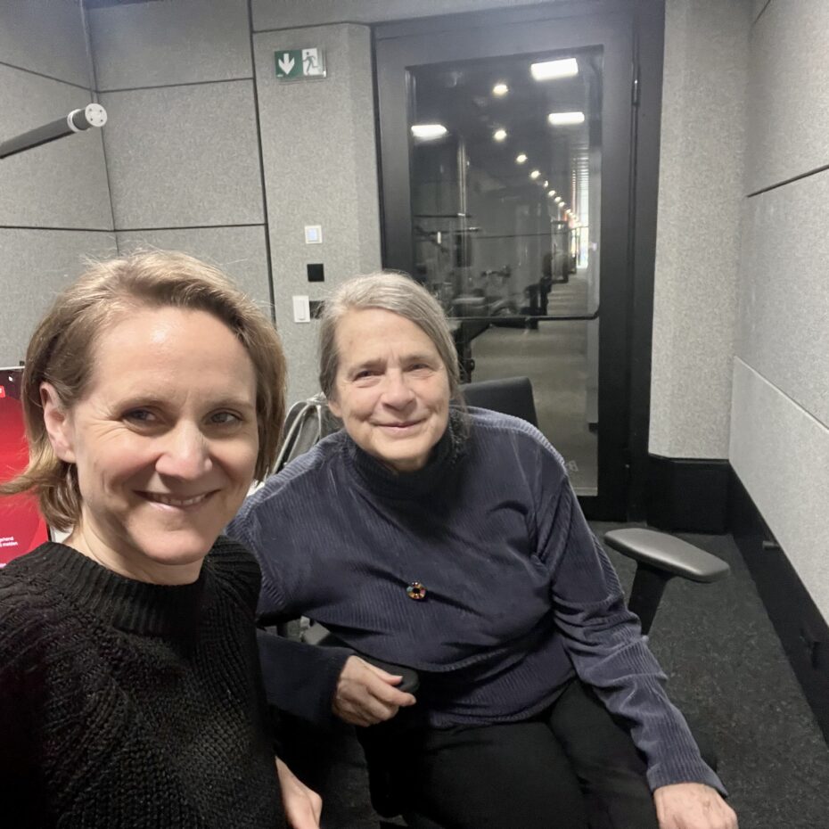 eine Frau mit grauen Haaren sitzt schräg hinter einer jüngeren Frau mit blonden Haaren in einem kleinen Tonstudio. Beide lächeln in die Kamera.