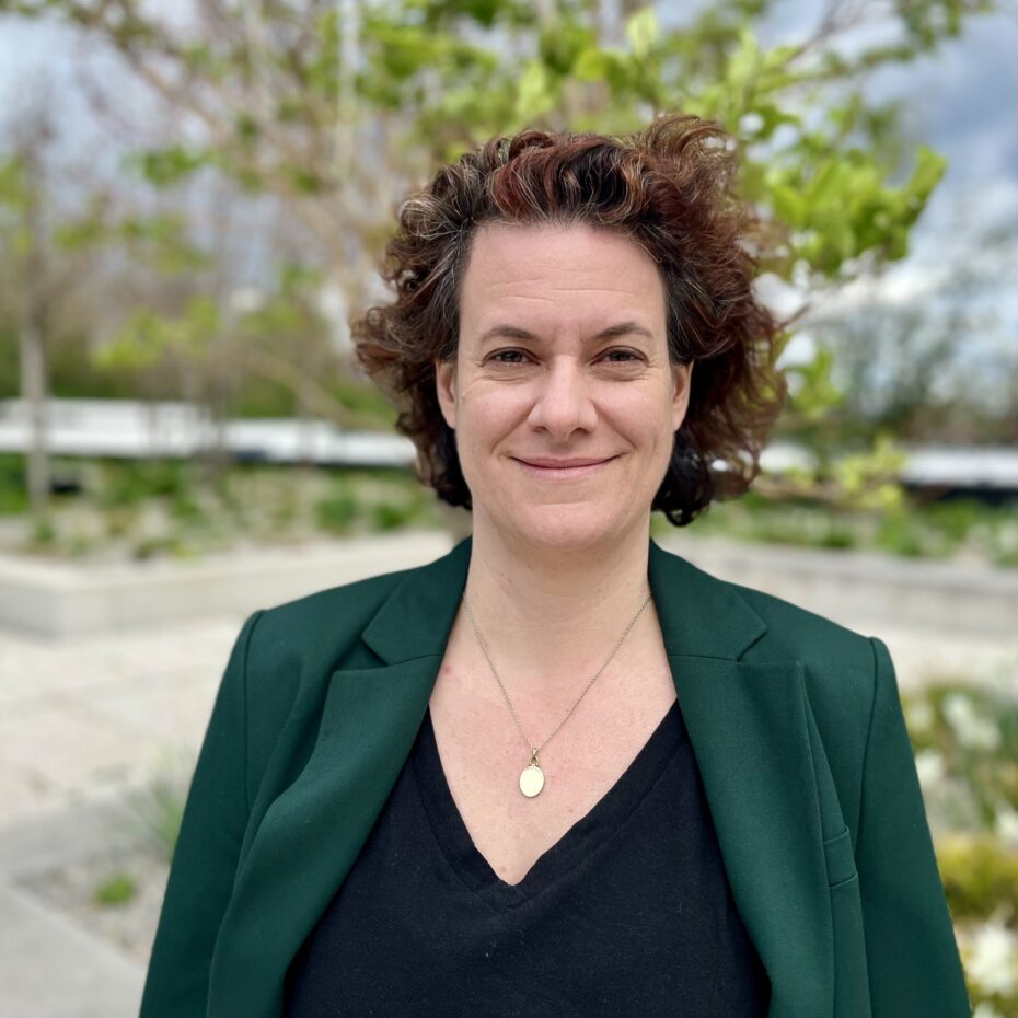 eien Frau mit kinnlangen Locken in dunkler Bluse und grünem Blazer lächelt in die Kamera. Der Wind spielt in ihren Haaren. Im Hintergrund sind zarte grüne Blätter an den Frühlingsbäumen in urbaner Landschaft zu erahnen.