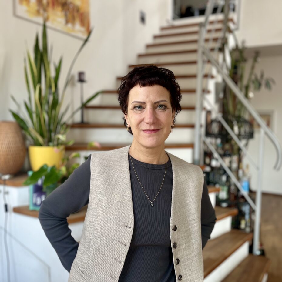 eine Frau mit kurzen dunklen Haaren lächelt in die Kamera. Im Bildhintergrund des Wohnzimmers führt eine Treppe hinauf.