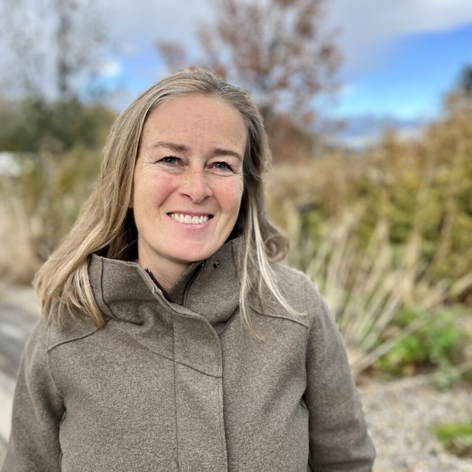 eine Frau mit blonden feinen Haaren, die ihr über die Schulter reichen. Sie trägt einen hellbraunen Mantel und steht vor einem grün-urbanen Hintergrund. Sie lächelt in die Kamera