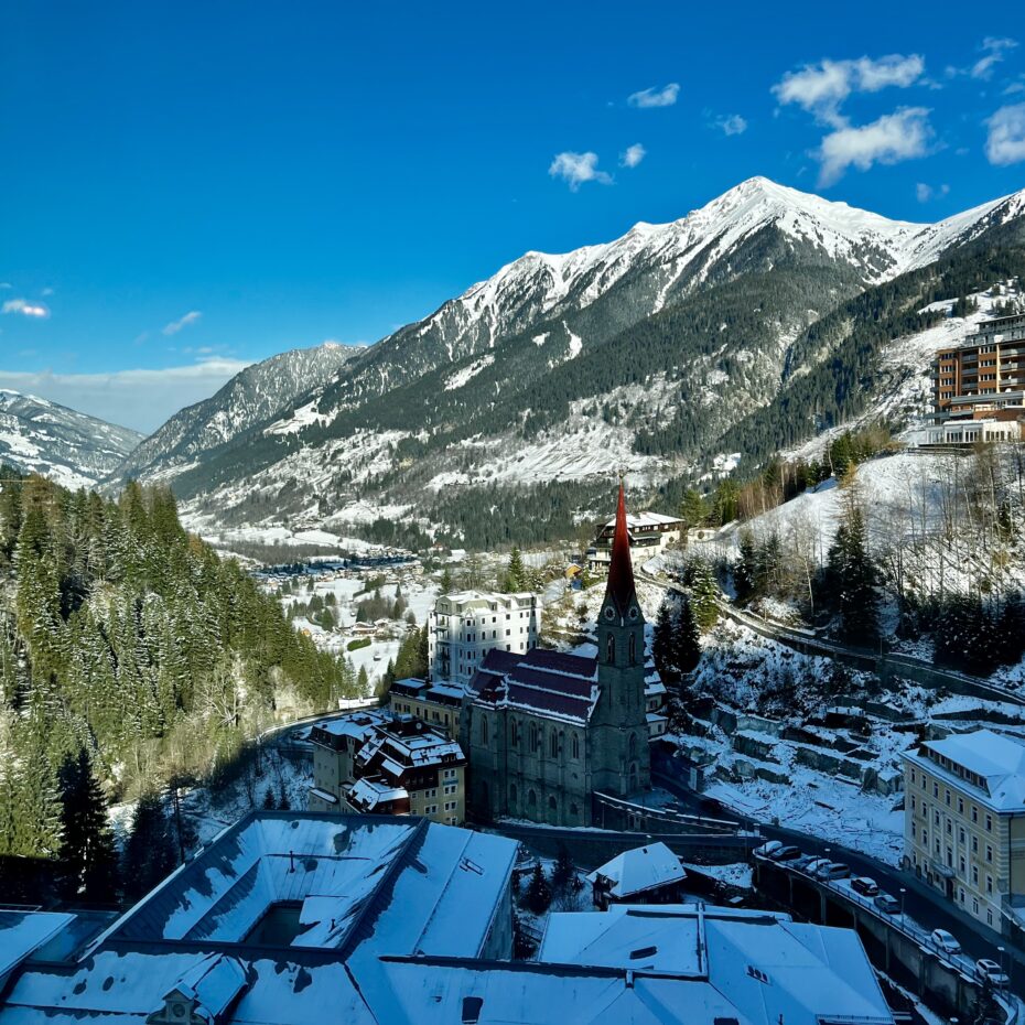 Blick von oben auf die Filialkirche St. Nikolaus. Die Kirche steht eng an den Hand geschmiegt inmitten von leicht schneebedeckten Häusern, die sich ebenfalls an den Hang in dem engen Talschluss schmiegen. Dahinter öffnet sich das Tal und der Blick auf angezuckerte Berge im Gasteiner Tal wird frei.