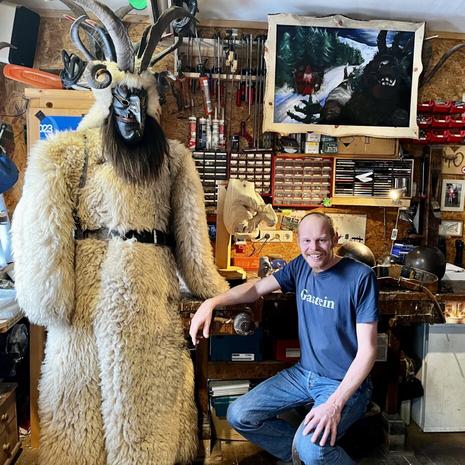 Ein Mann in Jeans und blauem T-Shirt mit der Aufschrift "Gastein" sitzt in einer Werkstatt neben einem mannsgroßen Krampus-Modell; d.h. einem Mantel aus Lammfell, auf dem ein schwarzer Krampuskopf mit Bart und Hörnern thront.