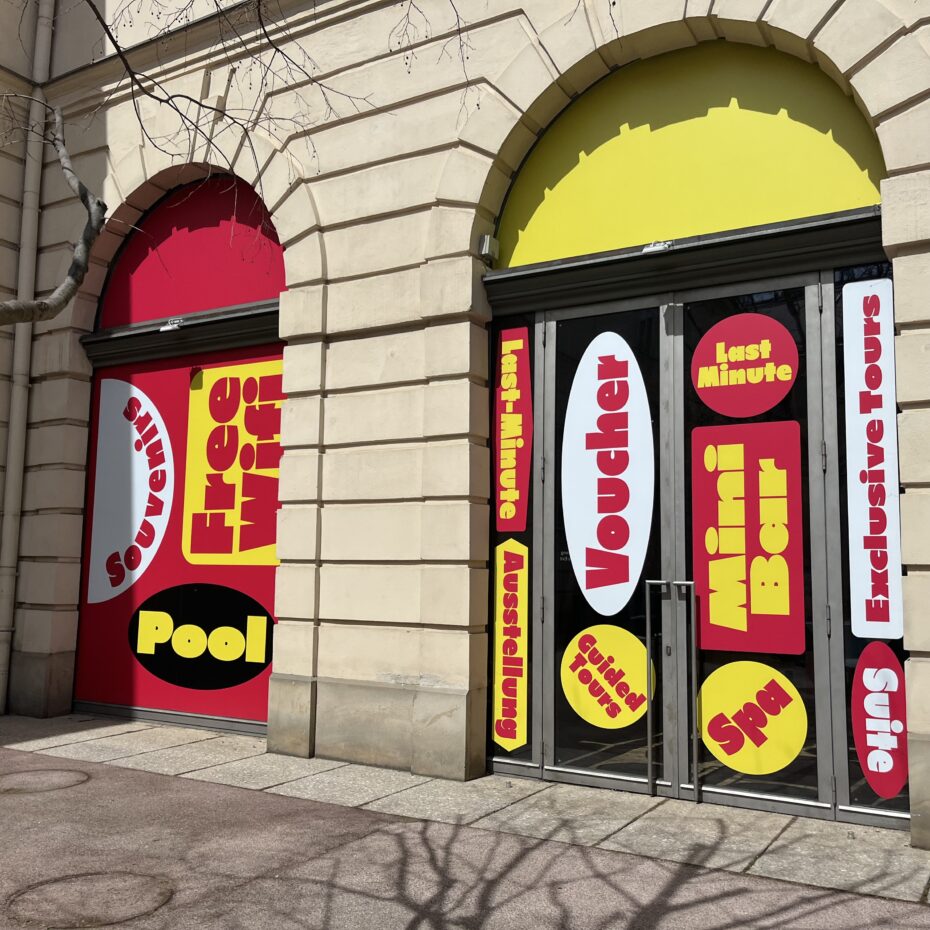 Auf zwei großen Schaufenstern kleben riesige neonfarbige Stikcer mit den Aufschriften: Pool, Voucher, Last-Minute, Spa, Guided Tours, Souvenirs, Free Wi-Fi, Mini Bar, u.a.
