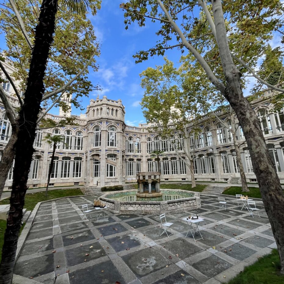 der weitläufige Innenhof eines Gebäudes aus Sandstein, mit großen Fenstern. In der Mitte des Hofes steht ein Springbrunnen. Am Rand stehen alte hohe Bäume.