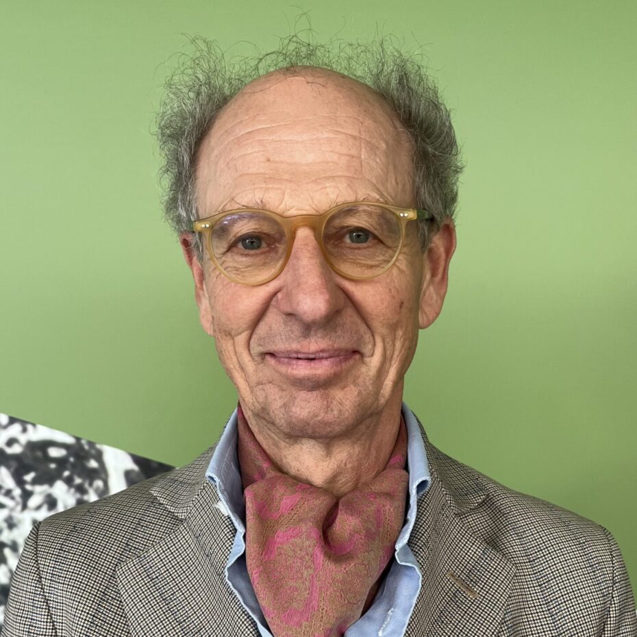 ein Mann mit gelockten Haaren rund um eine Halbglatzue, Brille und einem Sakko, in deren Brusttasche bunte Stifte stecken, stheht vor einer grünen Wand und lächelt in die Kamera