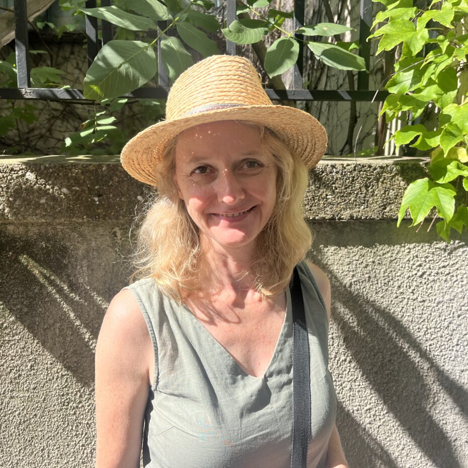 Eien blonde Frau mit Strohhut und einem Tank-Top steht im Schatten einer Art Hecke, die auf der Mauer hinter ihr wächst.