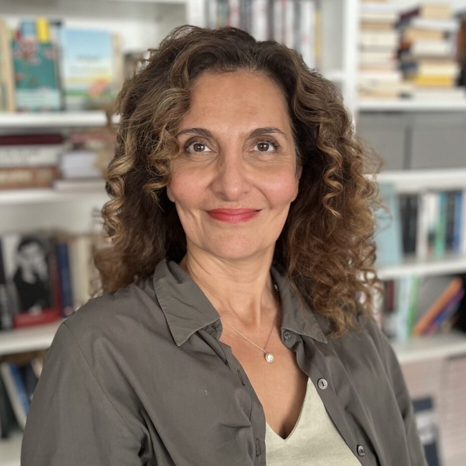 eine Frau mit dunklen Locken sitzt vor einem Bücherregal und blickt in die Kamera