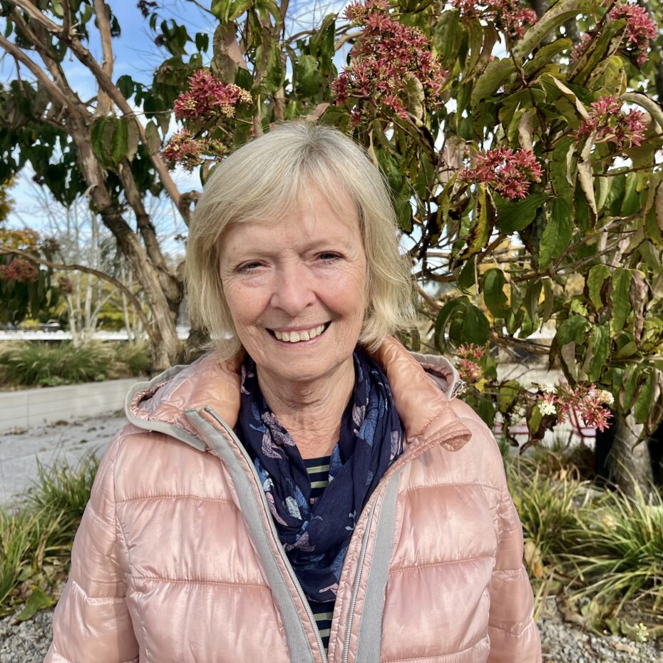eine Frau mit graublonden kinnlangen Haaren und einer rosa Daunenjacke steht vor einem Baum und blickt in die Kamera