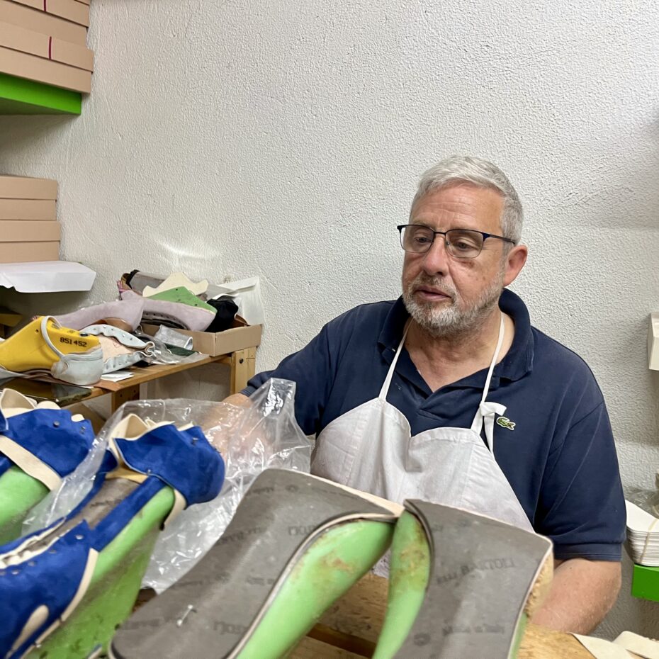 ein weißhaariger Mann mit Brille und einer Schürze sitzt umgeben von Damenschuhen mit Absätzen in verschiedenen Farben hinter seinem Arbeitstisch