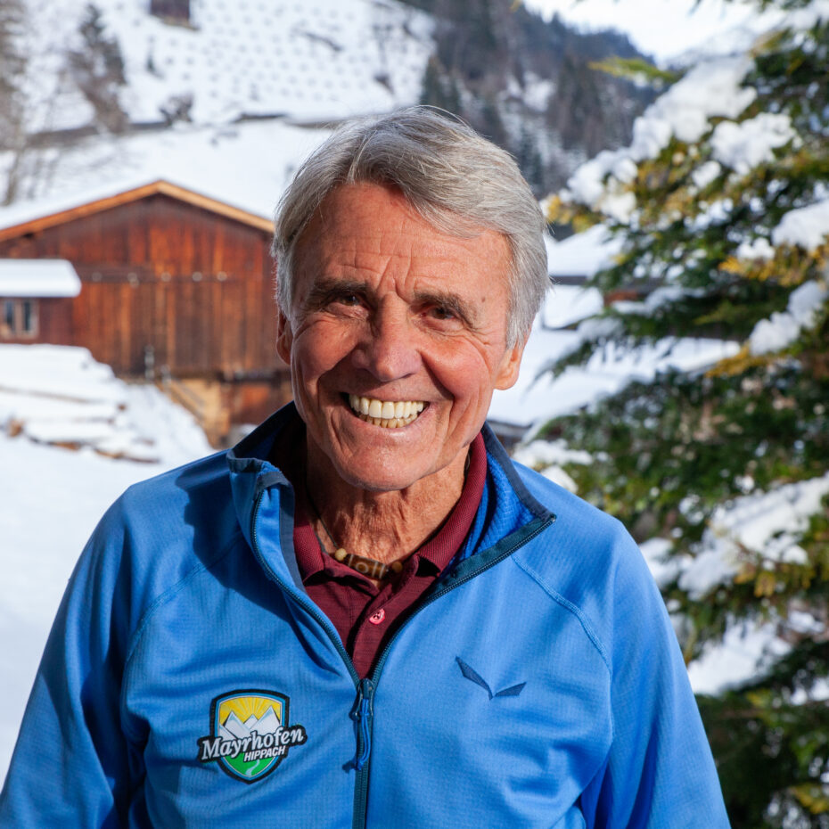 ein Mann mit grauem Haar steht vor einer verschneiten Winterlandschaft und strahlt lachend in die Kamera