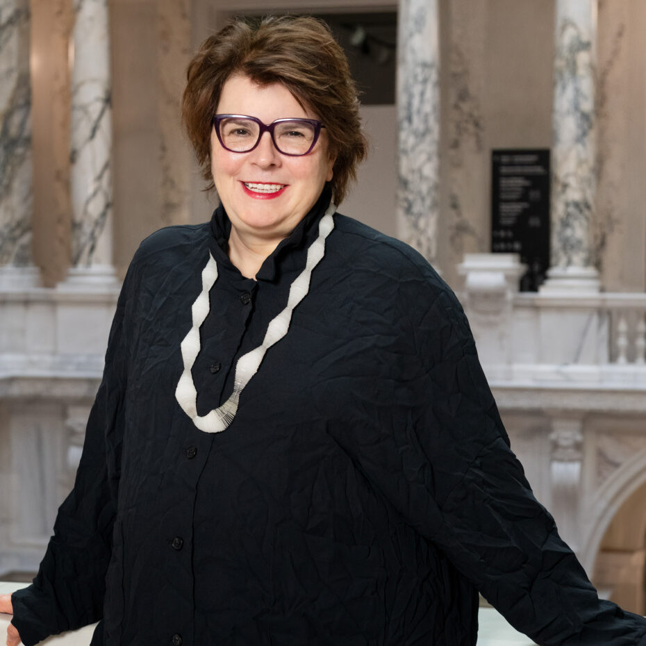 eine dunkelhaarige Frau mit Brille, schwarzem Pullover und einer großen Silberkette steht an einer Marmorbrüstung. Im Hintergrund Marmorsäulen und Brüstung auf der gegenüberliegenden Seite der Halle im Weltmuseum